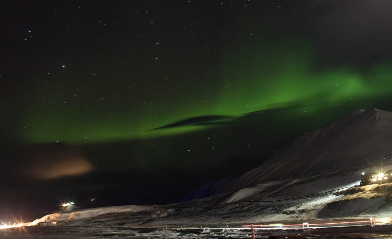 A green band of northern lights over a snowy mountain at the edge of Longyearbyen, with the town's lights glowing below