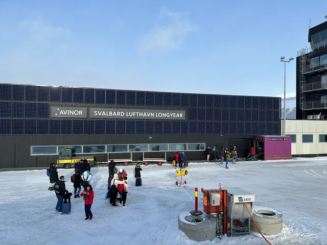 Passengers with suitcases walking across a snowy apron to the black terminal building marked Avinor, Svalbard Lufthavn Longyear