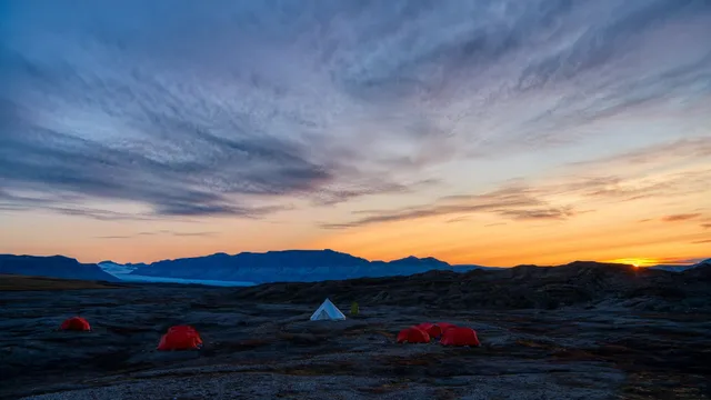 The midnight sun glowing orange on the horizon under streaked clouds, with a few red tents on a rocky plain in the foreground