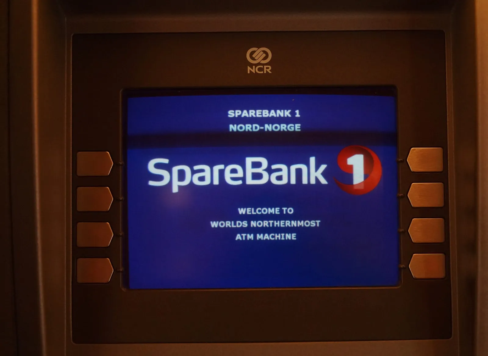 Der Bildschirm eines Geldautomaten in dunklem Rahmen mit dem Logo von SpareBank 1 und der Meldung „Welcome to world's northernmost ATM machine“