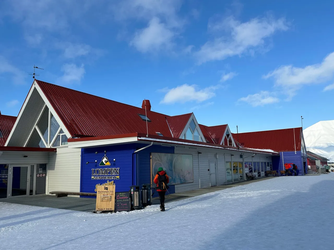 Das Einkaufszentrum Lompen in Longyearbyen, ein langes, niedriges Gebäude mit roten Dächern und blauem Eingang, an einem sonnigen Wintertag