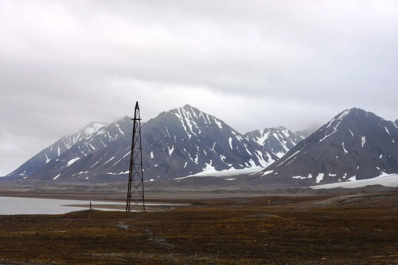 The tall, thin lattice airship mast standing alone on brown tundra in front of jagged, snow-streaked mountains