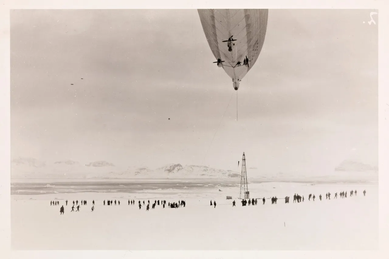 Black-and-white photo of the airship Norge hovering above a tall lattice mooring mast, with a line of people standing on the snow below