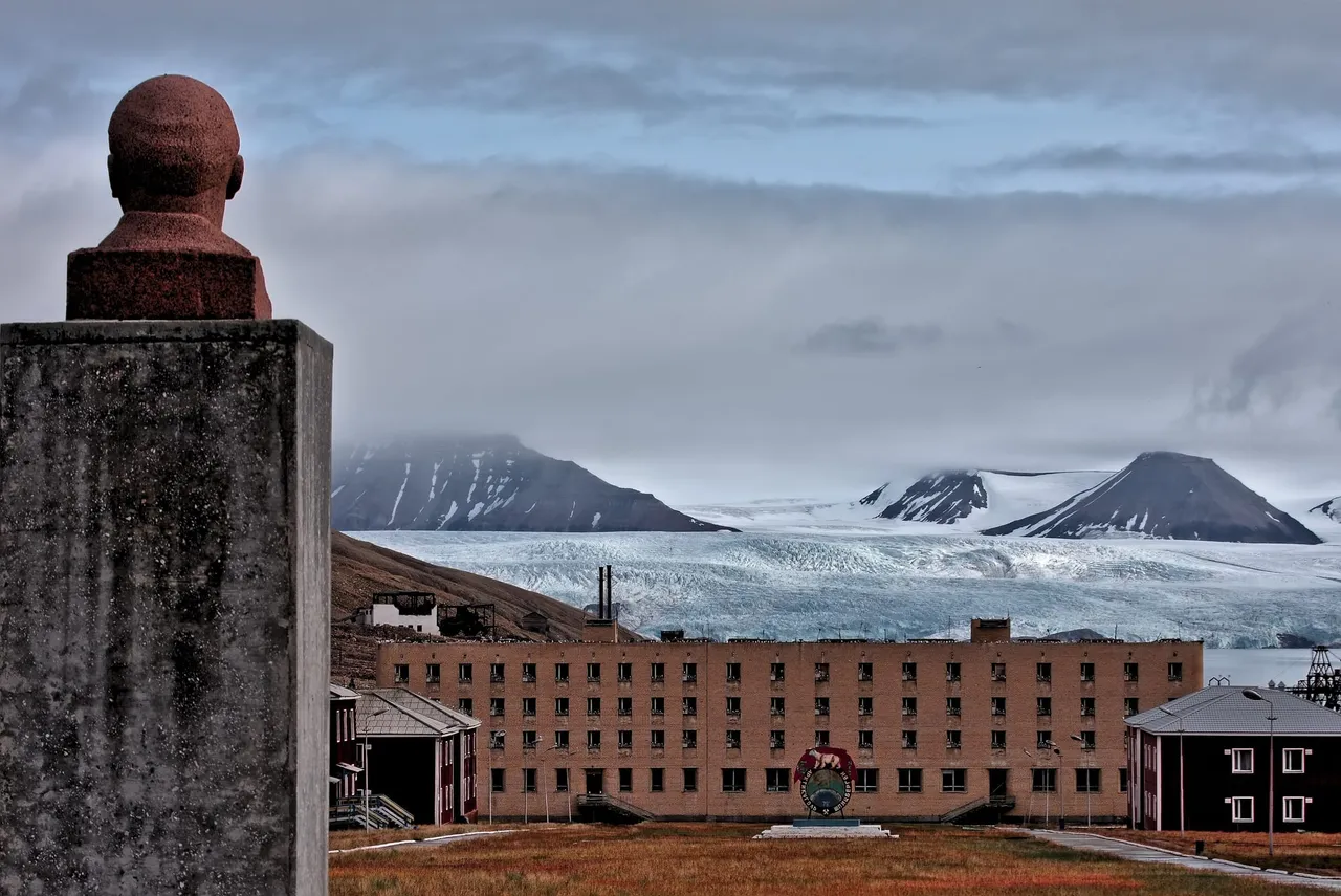 A dark stone bust of Lenin on a tall plinth in the foreground, with a long brick apartment block and the wide white front of a glacier behind