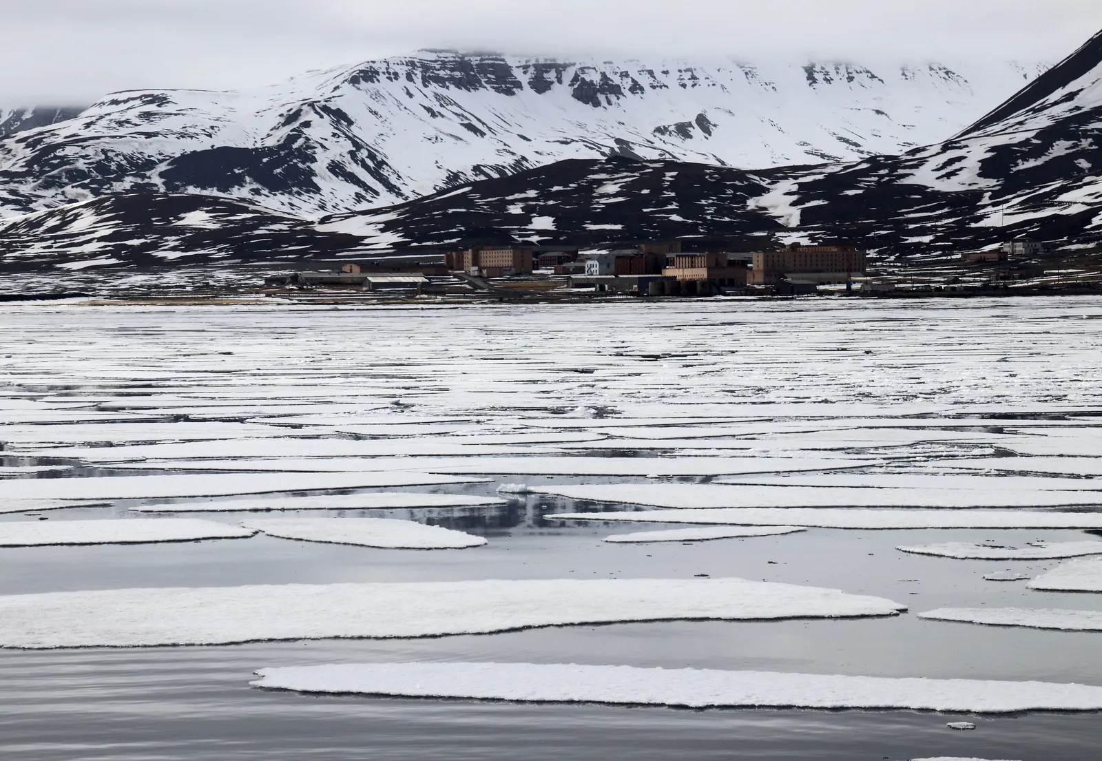 The brick buildings of Pyramiden seen in the distance across drifting sea ice, below dark, snow-streaked mountains